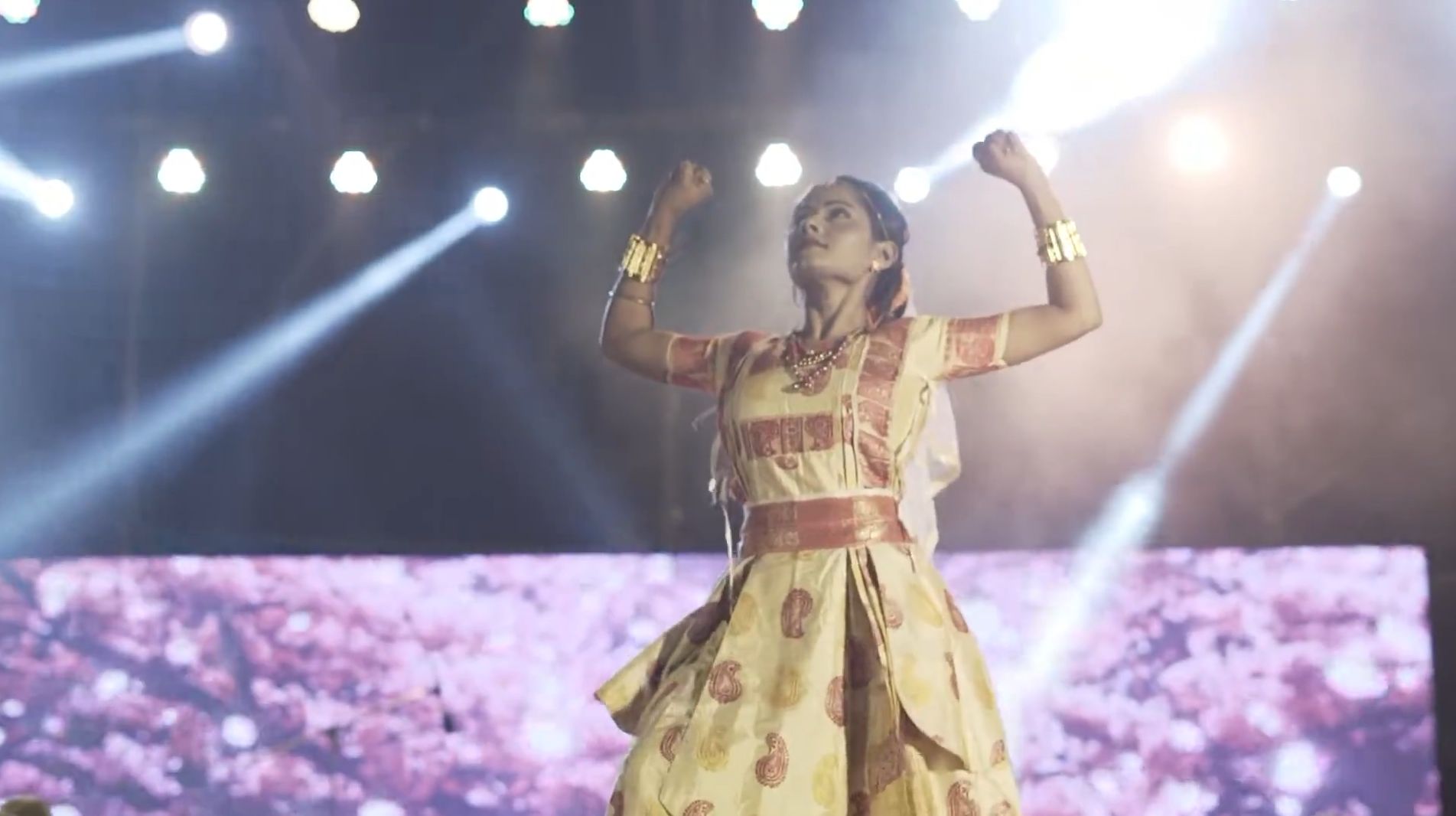 Still from the Cinematic Majuli Music Festival trailer. A dancer in a cream and red mekhela sador raises both arms mid-performance, lit from behind by stage spotlights.