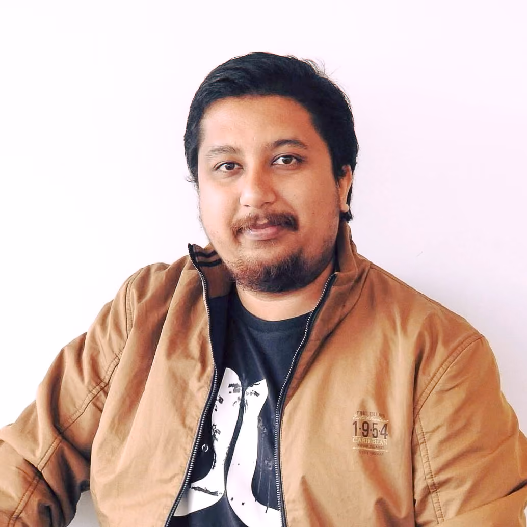 Portrait of filmmaker Kaushik Sharma in a tan jacket, seated against a plain white wall, looking towards the camera.