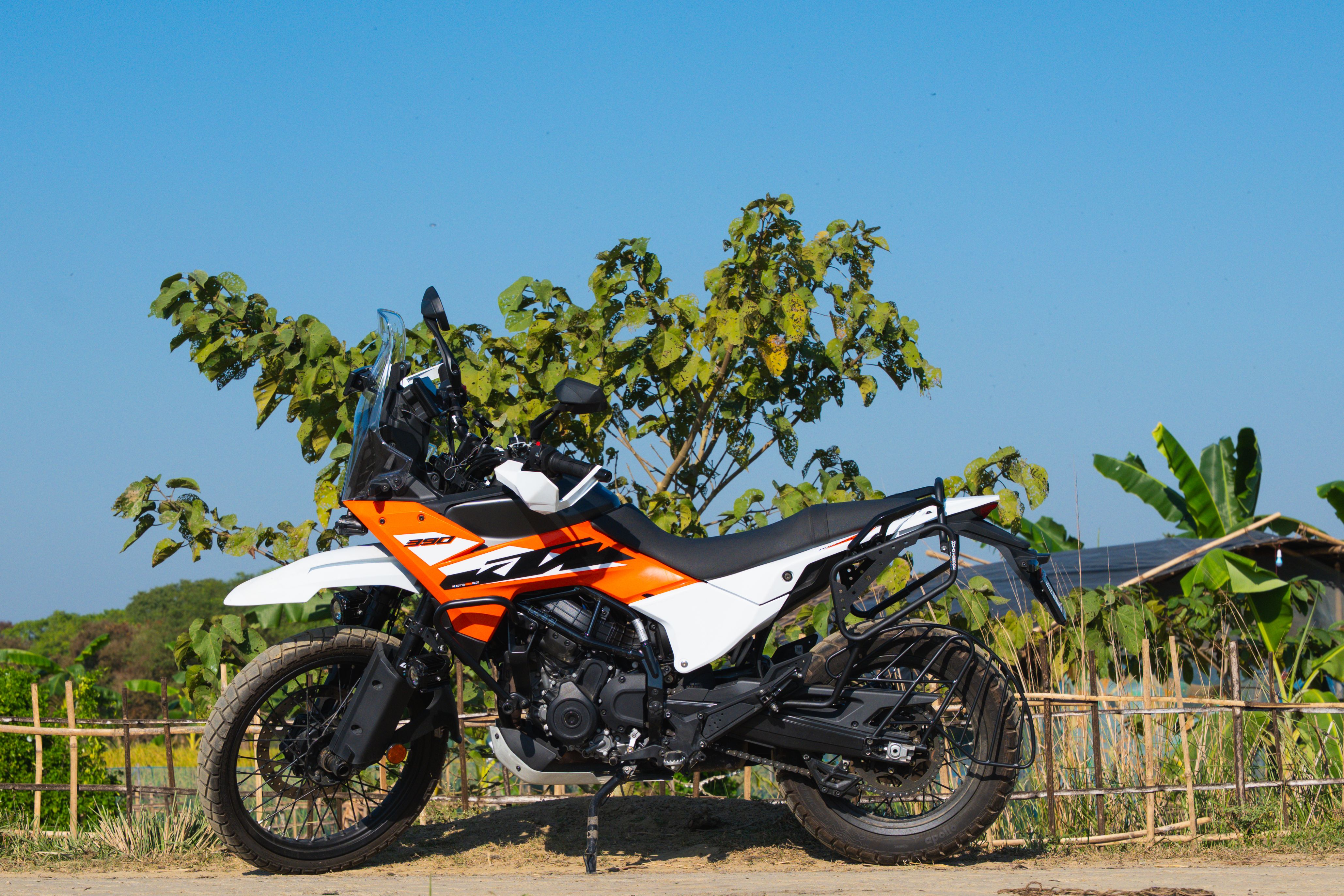 Kaushik Sharma’s orange and white KTM 390 Adventure parked on a dirt track beside a bamboo fence and banana plants under a clear blue Assam sky.