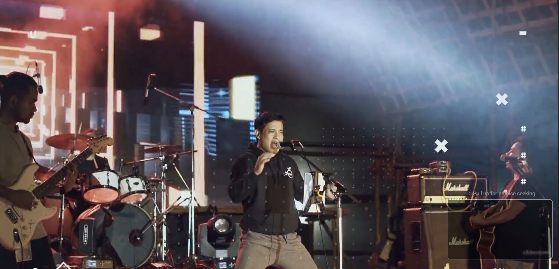 Still from the Majuli Music Festival promo. A singer performs mid-song at a microphone under a beam of stage light, flanked by a guitarist and a drum kit.
