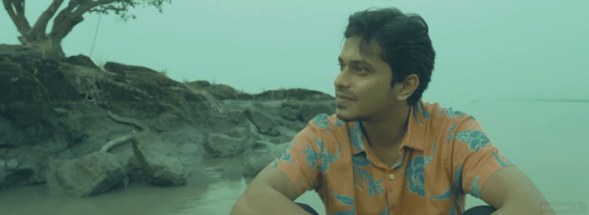 Still from The Scroll That Stole Time. Aarav sits on a riverbank in Guwahati at dusk in a patterned shirt, looking away from his phone across the water, in a cool teal grade.