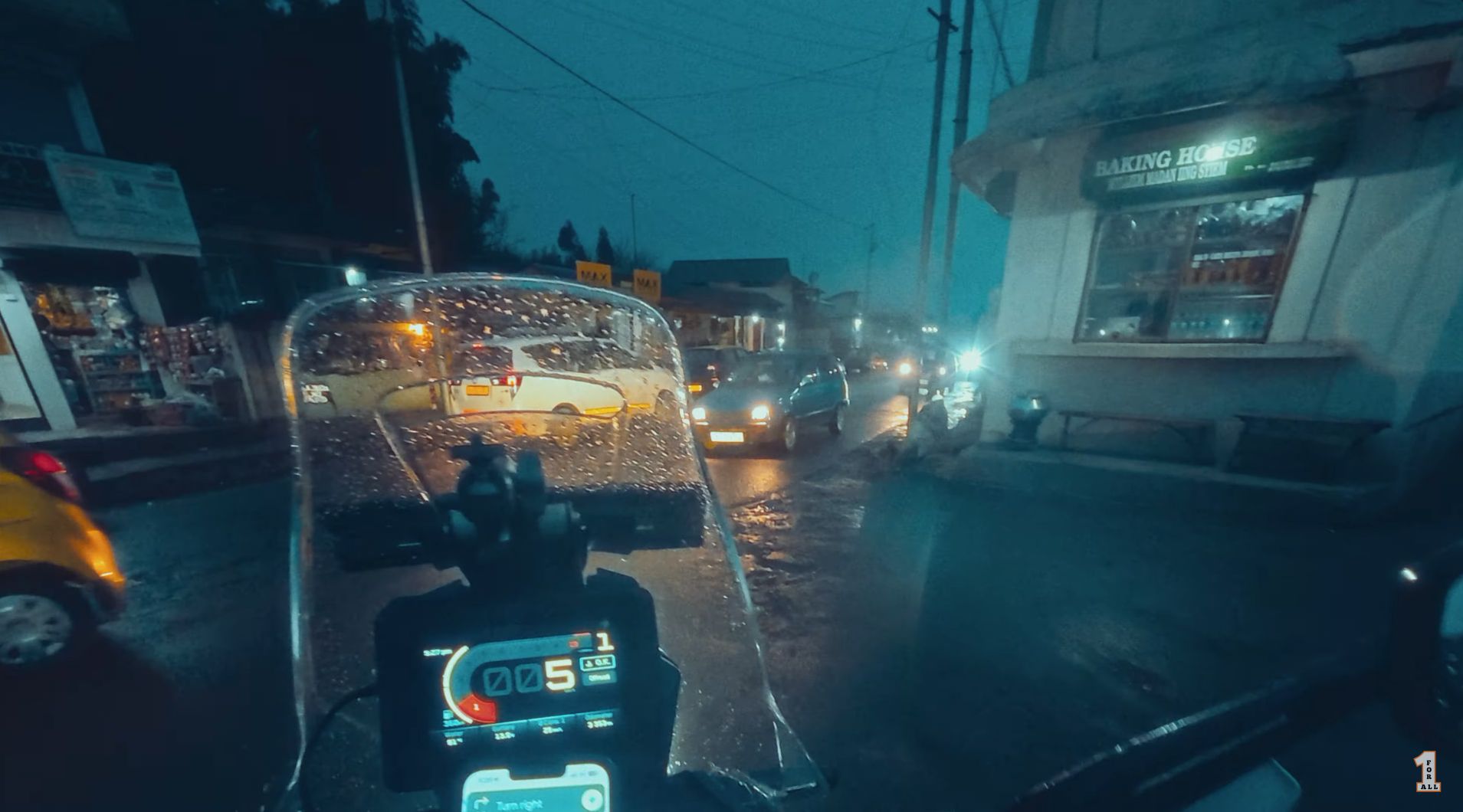 Rider’s-eye still from the Shillong to Cherrapunji motovlog. Handlebars and instrument cluster in the foreground, rain-slicked Meghalaya street traffic and shopfronts glowing blue at dusk ahead.
